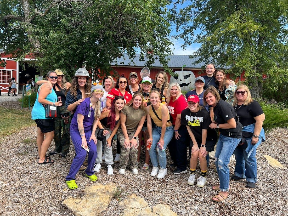 Group of smiling people posing outdoors on gravel in front of a red building and trees