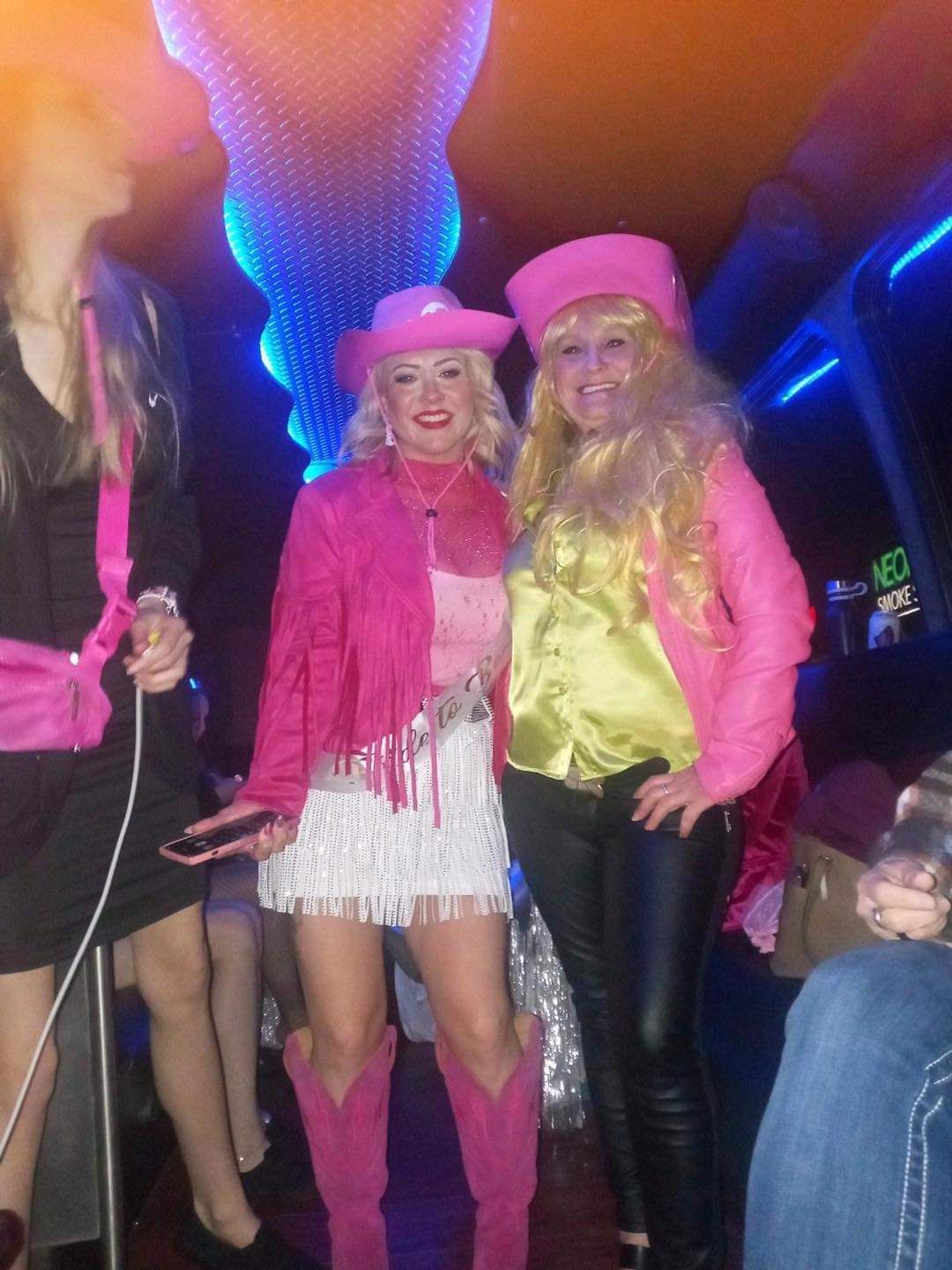 Two people in bright pink hats and jackets posing at a lively club with blue lighting