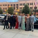 Group of people posing outdoors on a city sidewalk in formal dresses and suits at dusk.