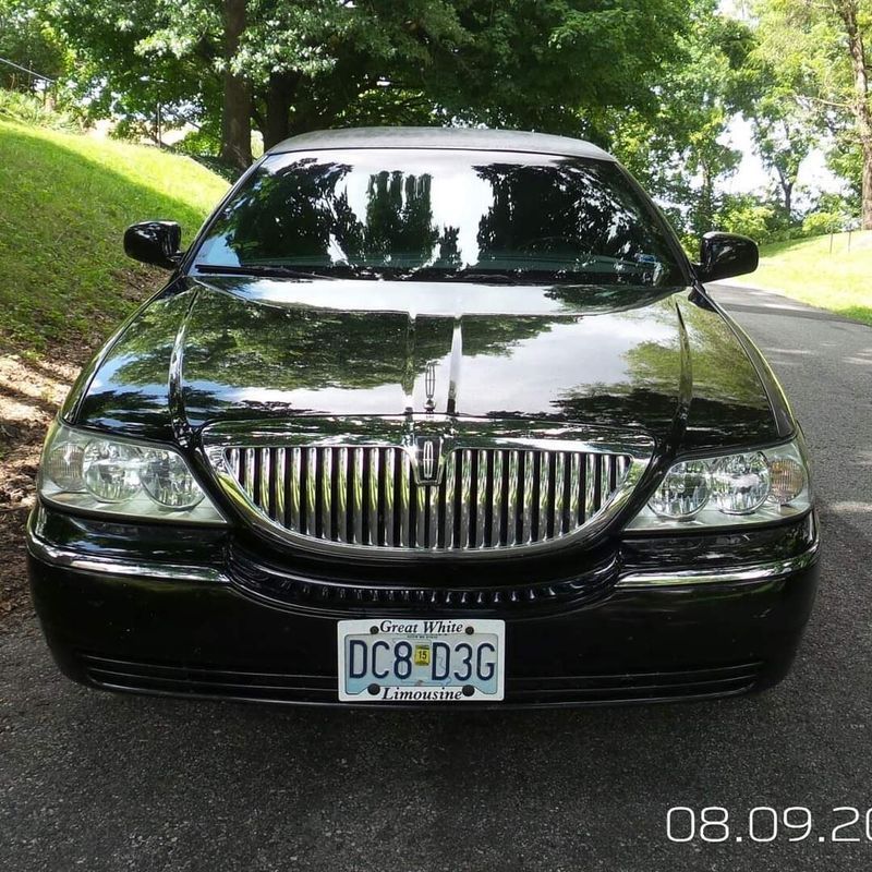 Black sedan parked on a tree-lined road, front view with chrome grille and license plate.