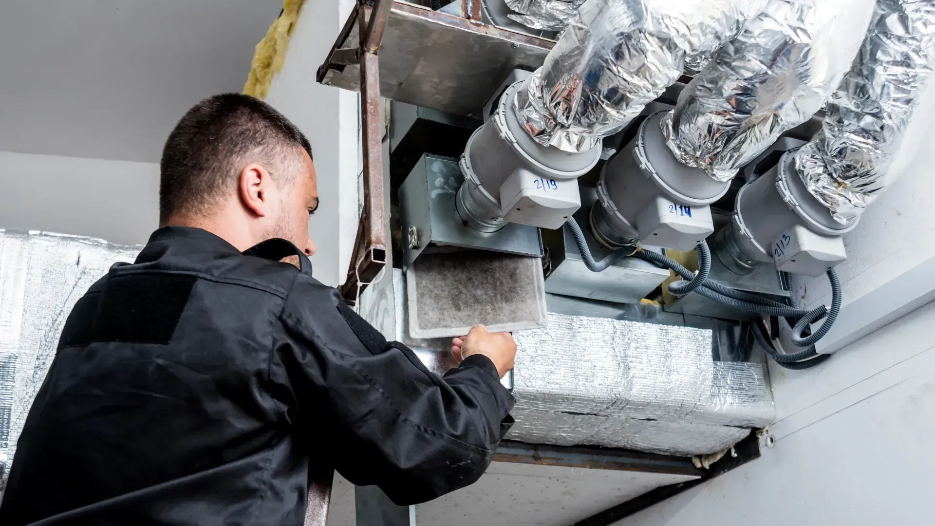 A man in black work clothes inspects HVAC ductwork, under a bright light.