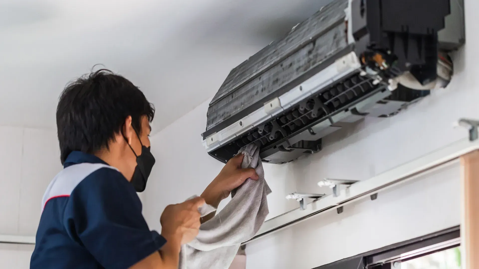 Person in a dark uniform cleaning a wall-mounted air conditioner with a white cloth, indoors.