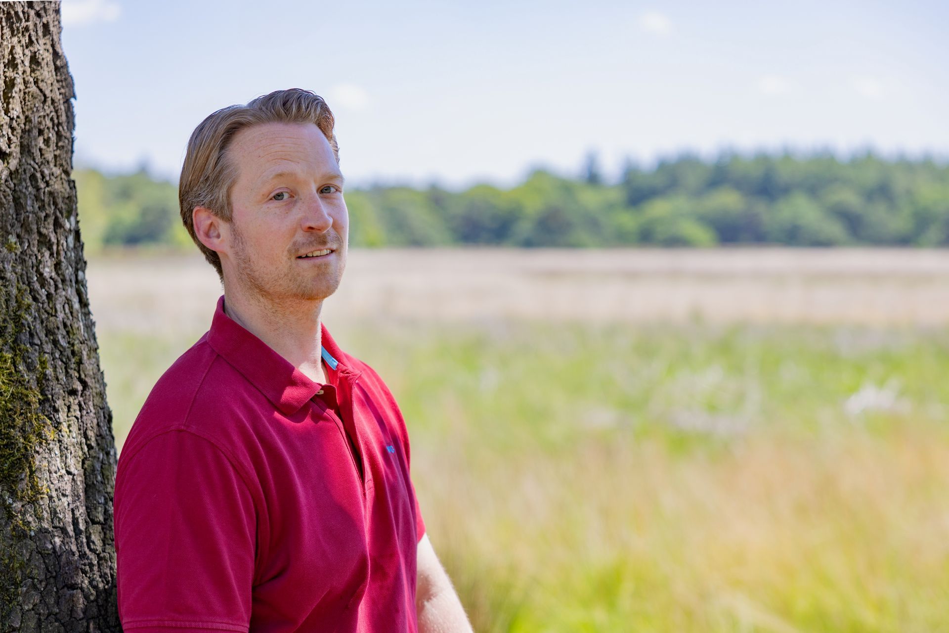 Een man in een rood shirt staat bij een boom in een zonnig veld met gras en wazige bomen op de achtergrond.