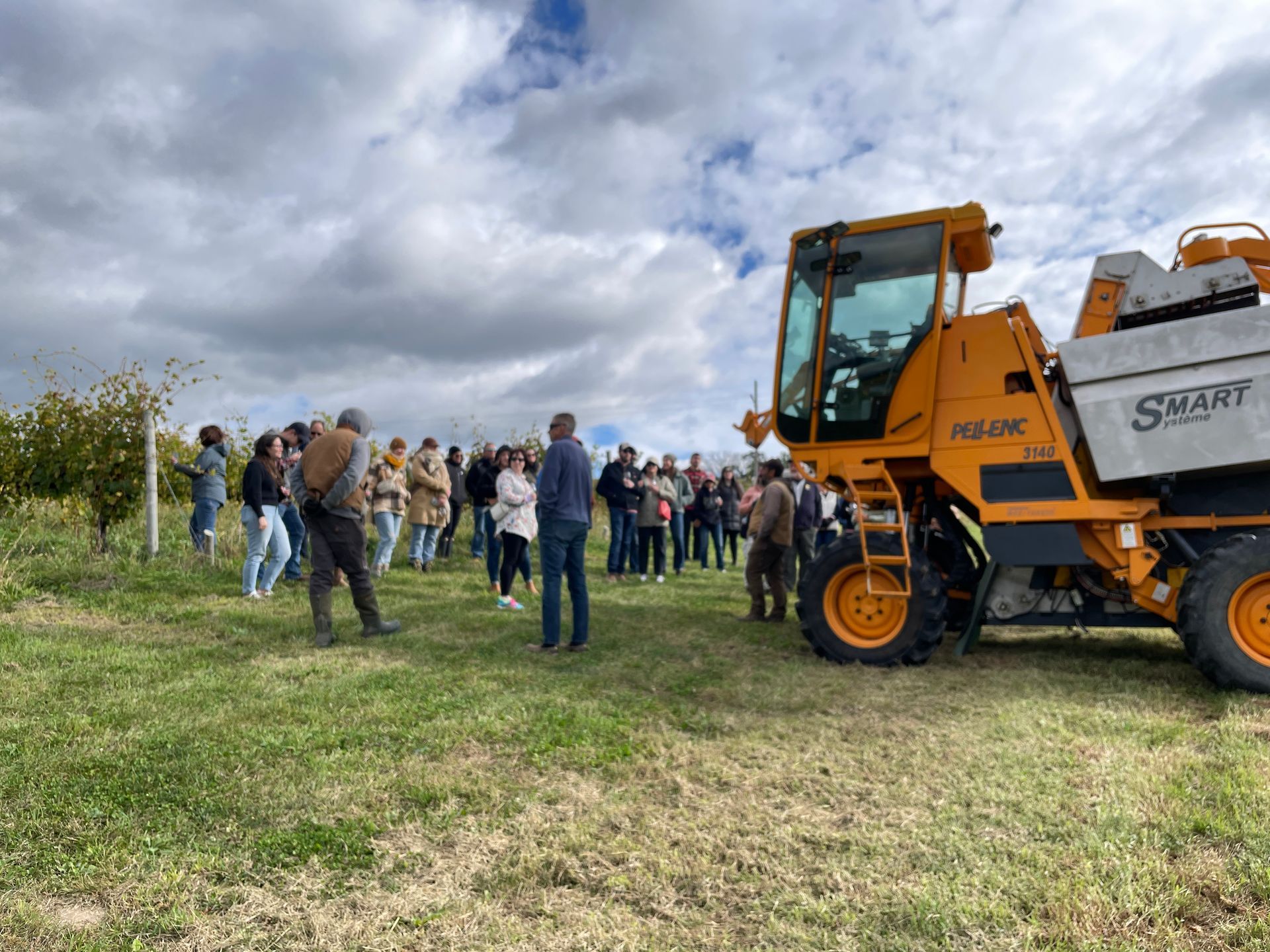 Group of people in a vineyard, listening to someone near an orange harvesting machine on a cloudy day.
