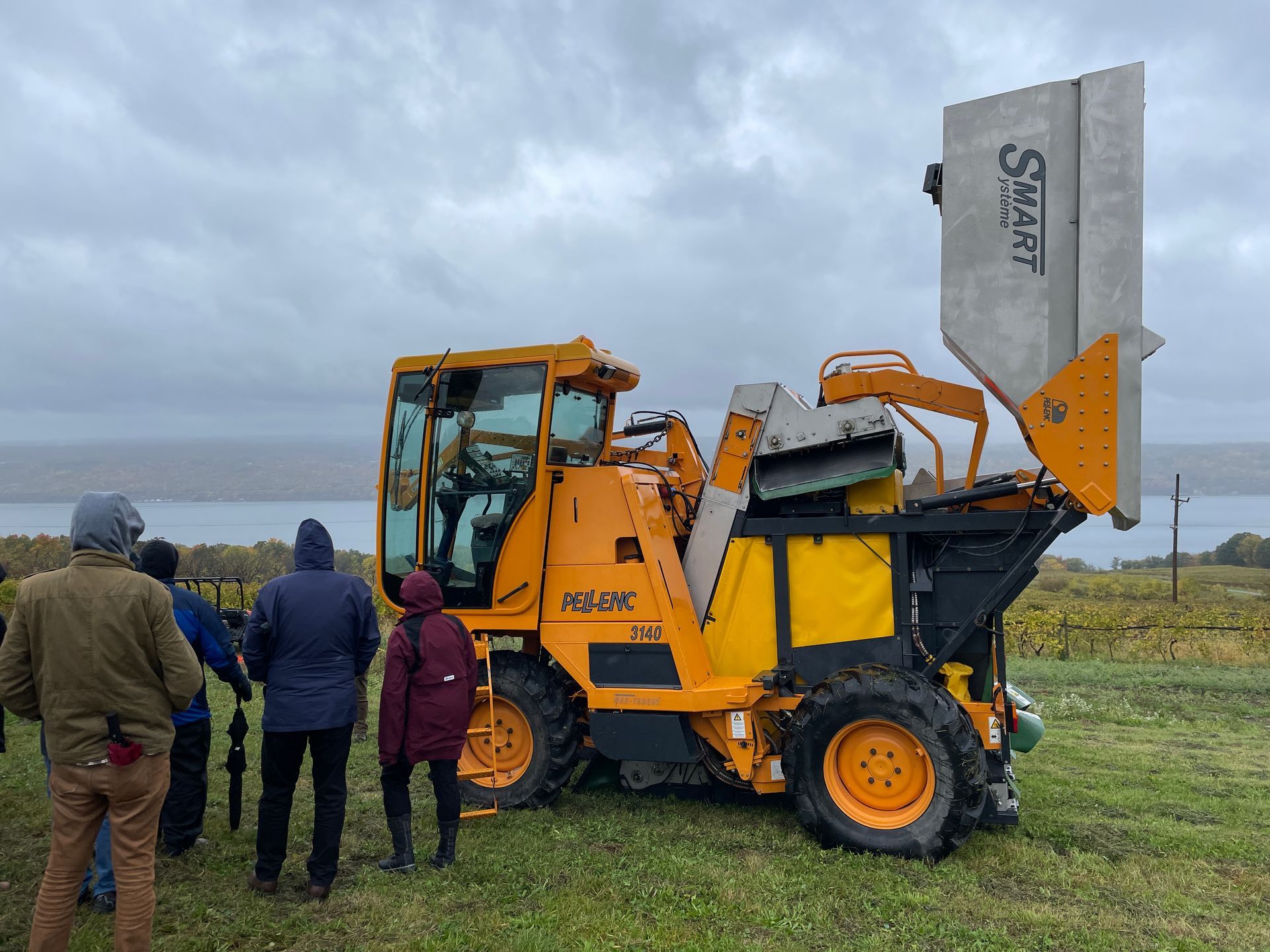 People inspecting a yellow agricultural machine with a raised white hopper in a field under cloudy skies