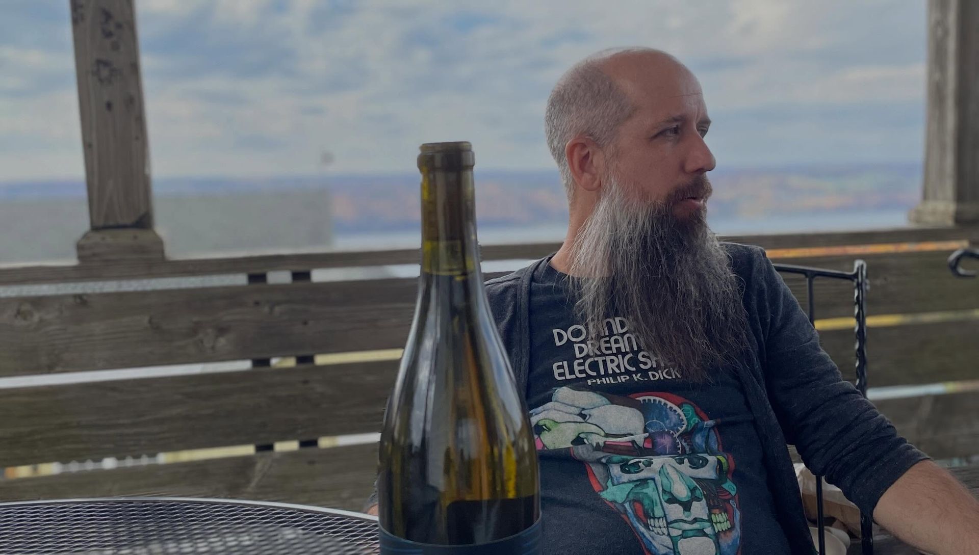 Man seated on a deck, looking right, with a bottle on a table in front and mountains in the background