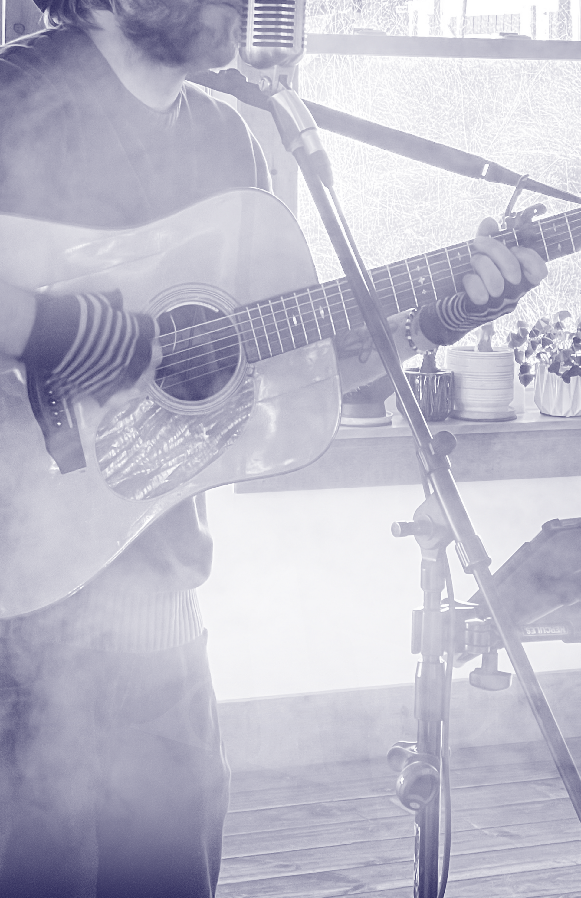 A musician plays an acoustic guitar and sings into a vintage-style microphone on a stand in a brightly lit room.