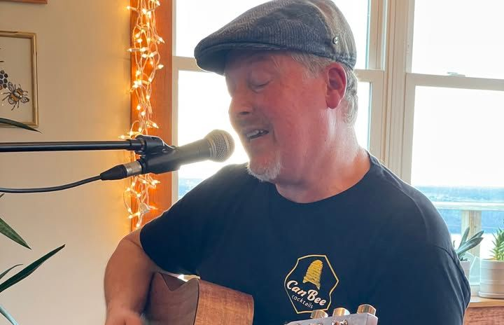 A person wearing a flat cap sings into a microphone while playing an acoustic guitar indoors near a window with lights.