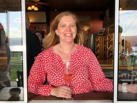 Woman in red shirt smiles, holding a pink drink, framed by a window, possibly a bar or restaurant setting.