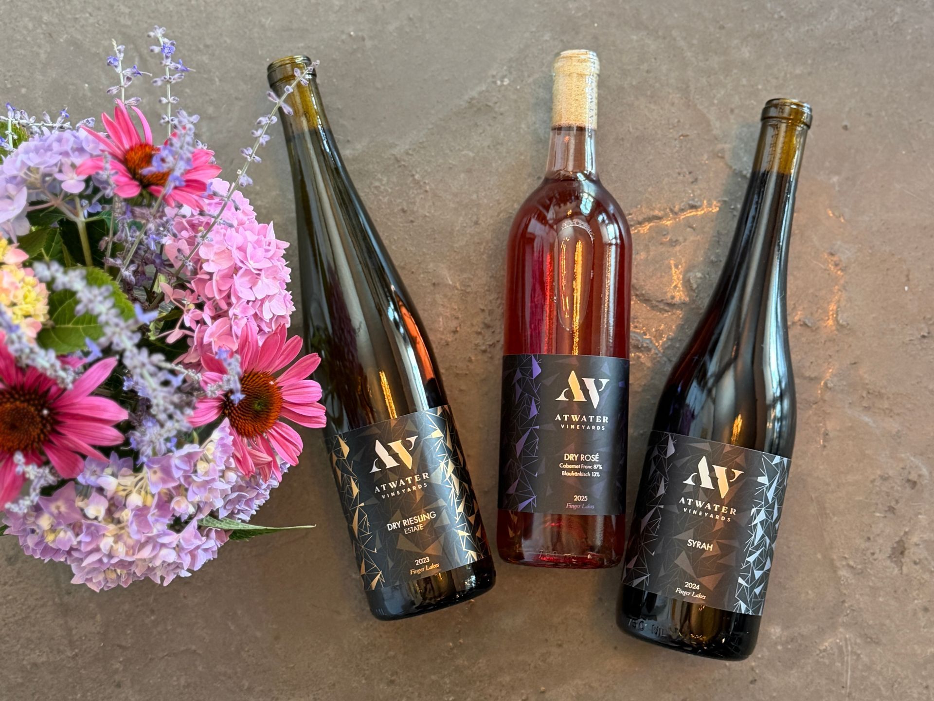 Three wine bottles beside a bouquet of pink and purple flowers on a gray surface