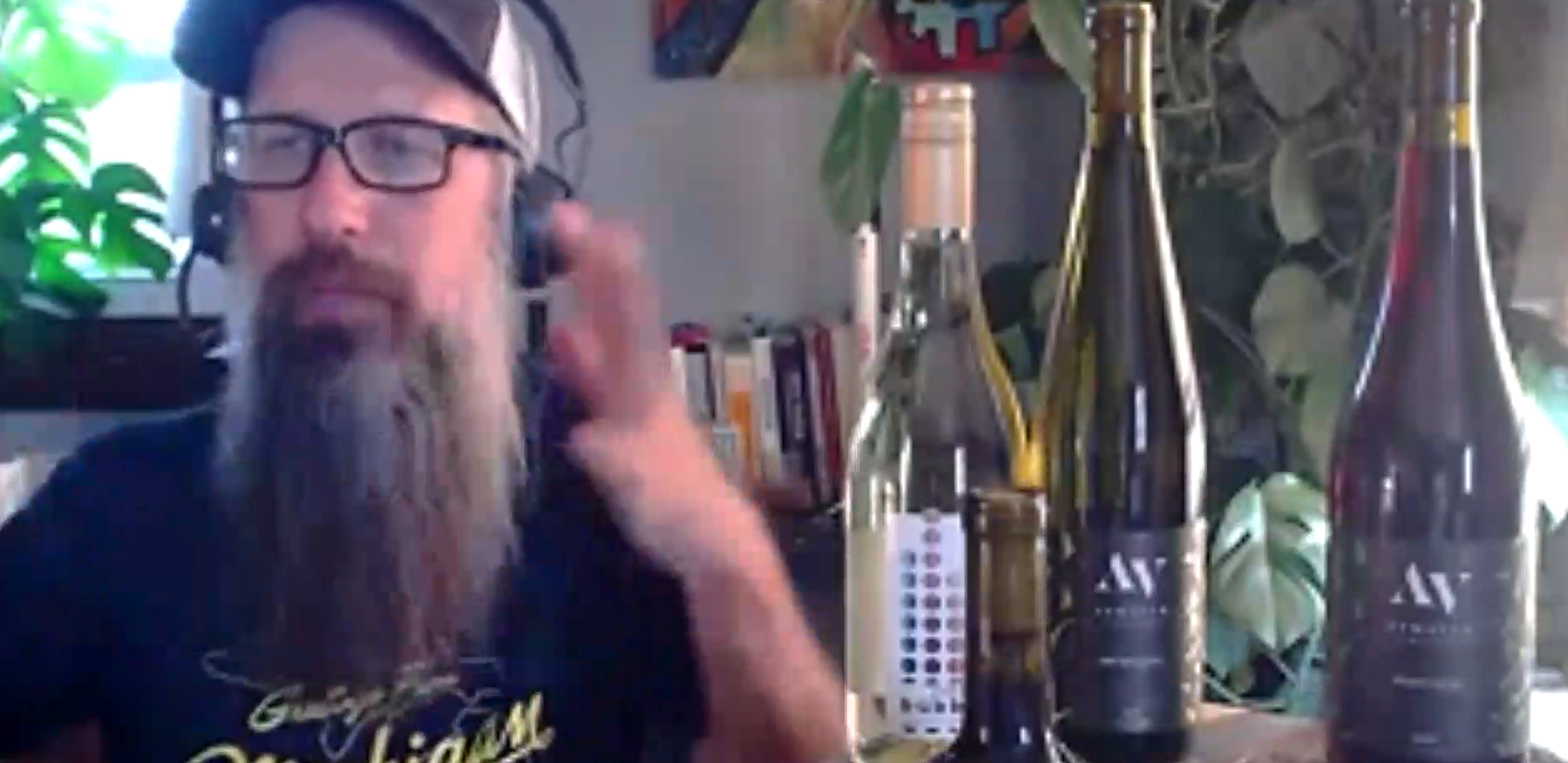 Man with glasses and long beard speaking beside several wine bottles on a table