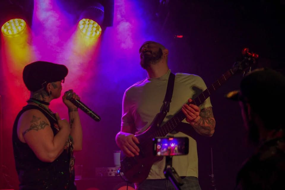 Two musicians performing on a purple-lit stage, one singing into a mic while another plays guitar.