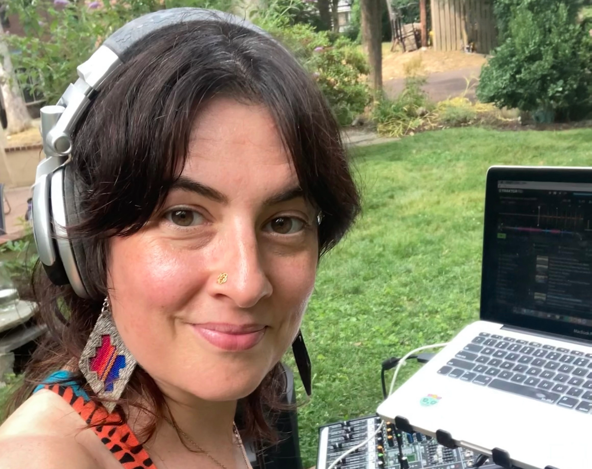 Smiling person wearing headphones beside a laptop outdoors in a green garden