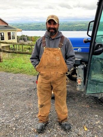 Man in brown overalls and gray hoodie standing near machinery, overlooking a body of water.