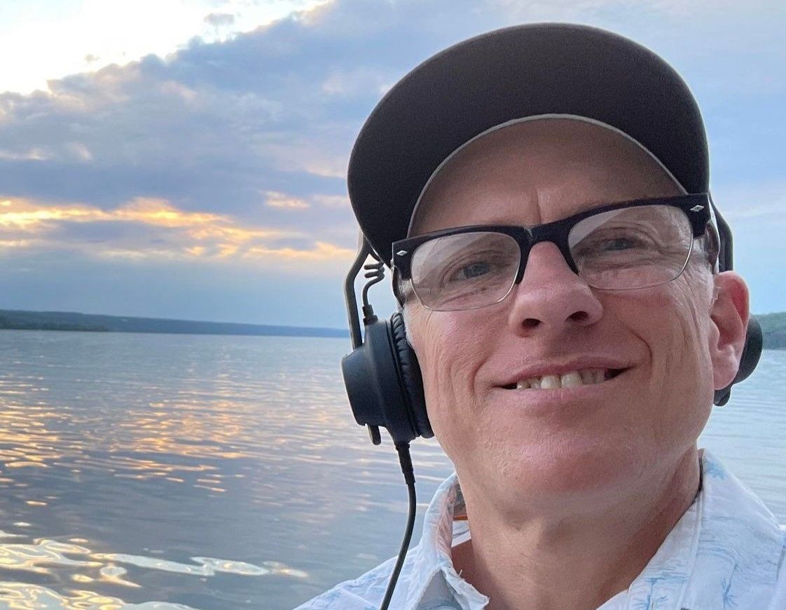 Smiling person in glasses and cap wearing headphones, standing by a calm waterfront at sunset