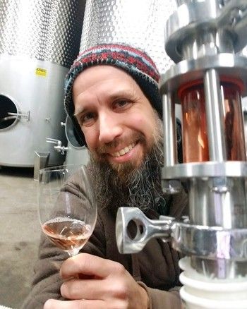 Man with a beard, holding rosé, smiling in a winery. Stainless steel tanks and equipment in the background.