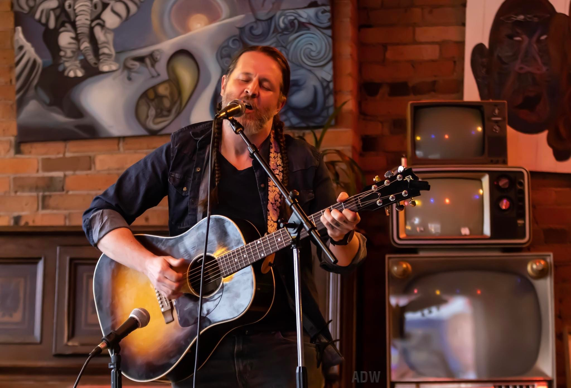 Singer playing acoustic guitar onstage in a brick-walled room with retro TVs behind him
