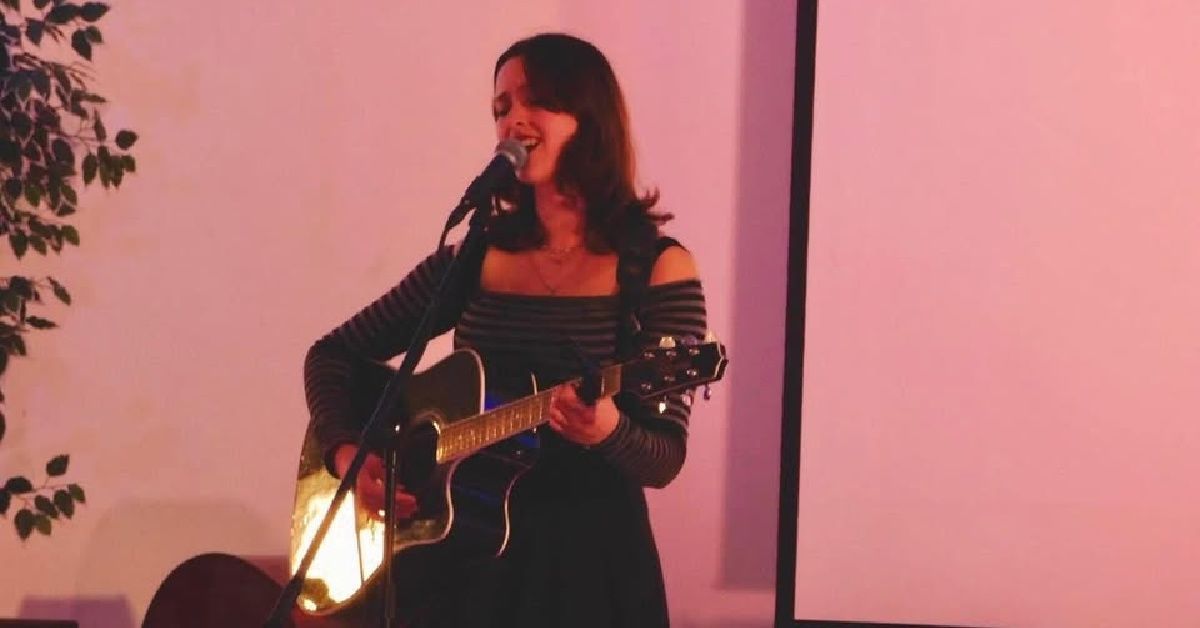 Woman singing and playing guitar on stage under pink lighting