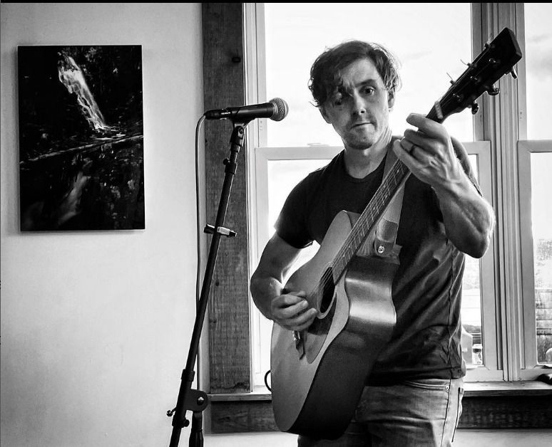A person with a neutral expression plays an acoustic guitar next to a microphone stand in a room with a window.
