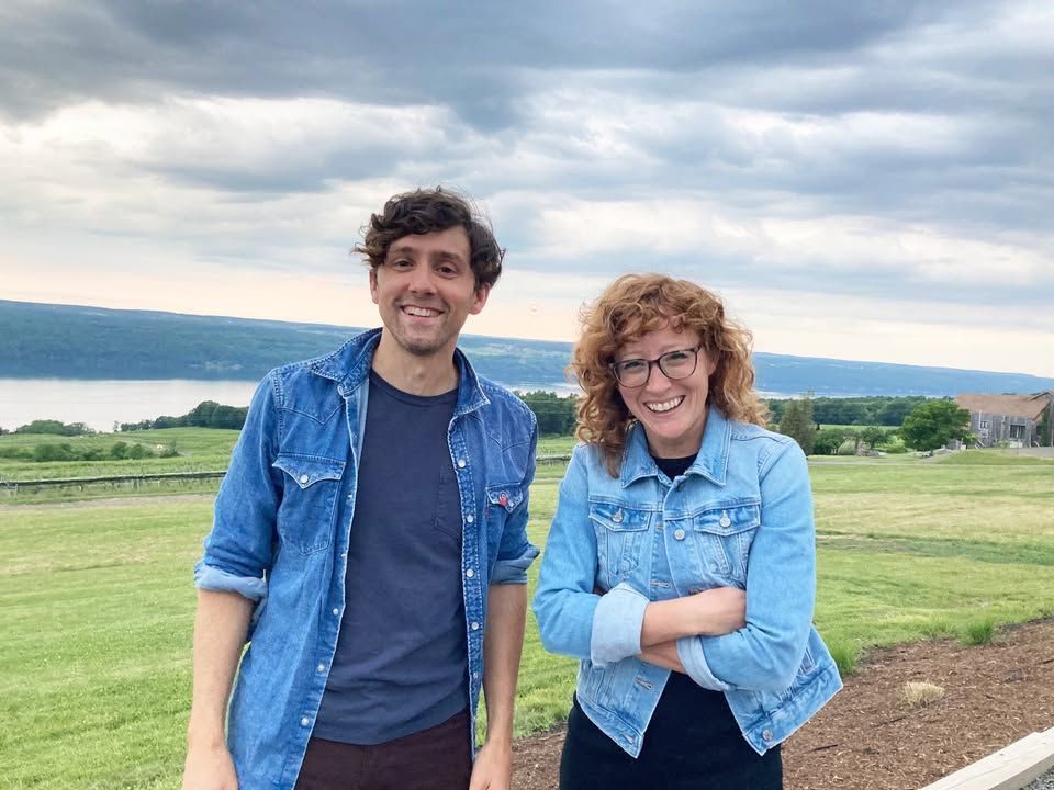 Two smiling people in denim jackets, posing outdoors with a lake and cloudy sky in the background.