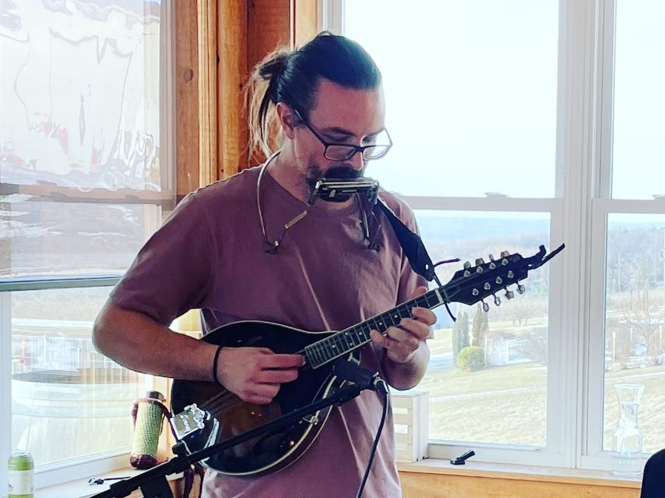 A person with a ponytail and glasses playing a mandolin and harmonica in front of a bright window.