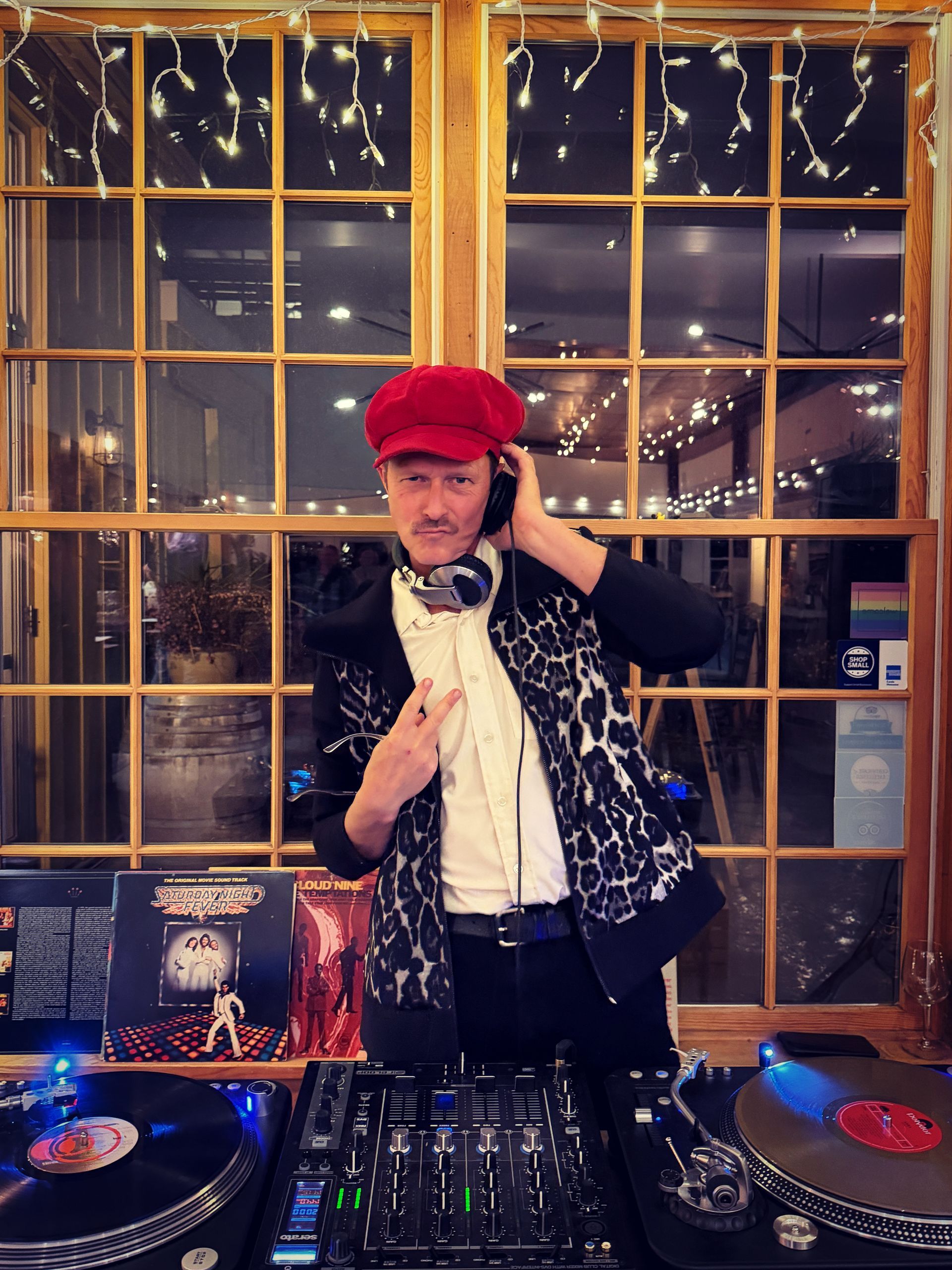 DJ wearing red hat and leopard jacket, smiling, gesturing at a DJ setup in front of a window.