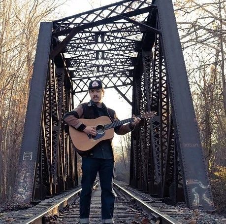Man standing with acoustic guitar on a steel railroad bridge, surrounded by bare trees.
