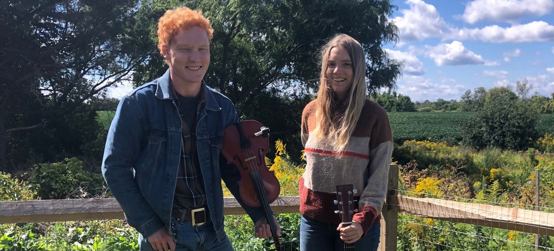 Two people standing by a wooden fence in a sunny field, one holding a guitar, with trees in the background