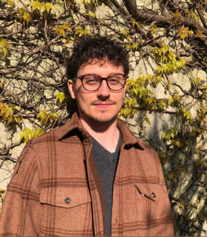 Man with glasses and brown plaid jacket smiles in front of a wall with vines.