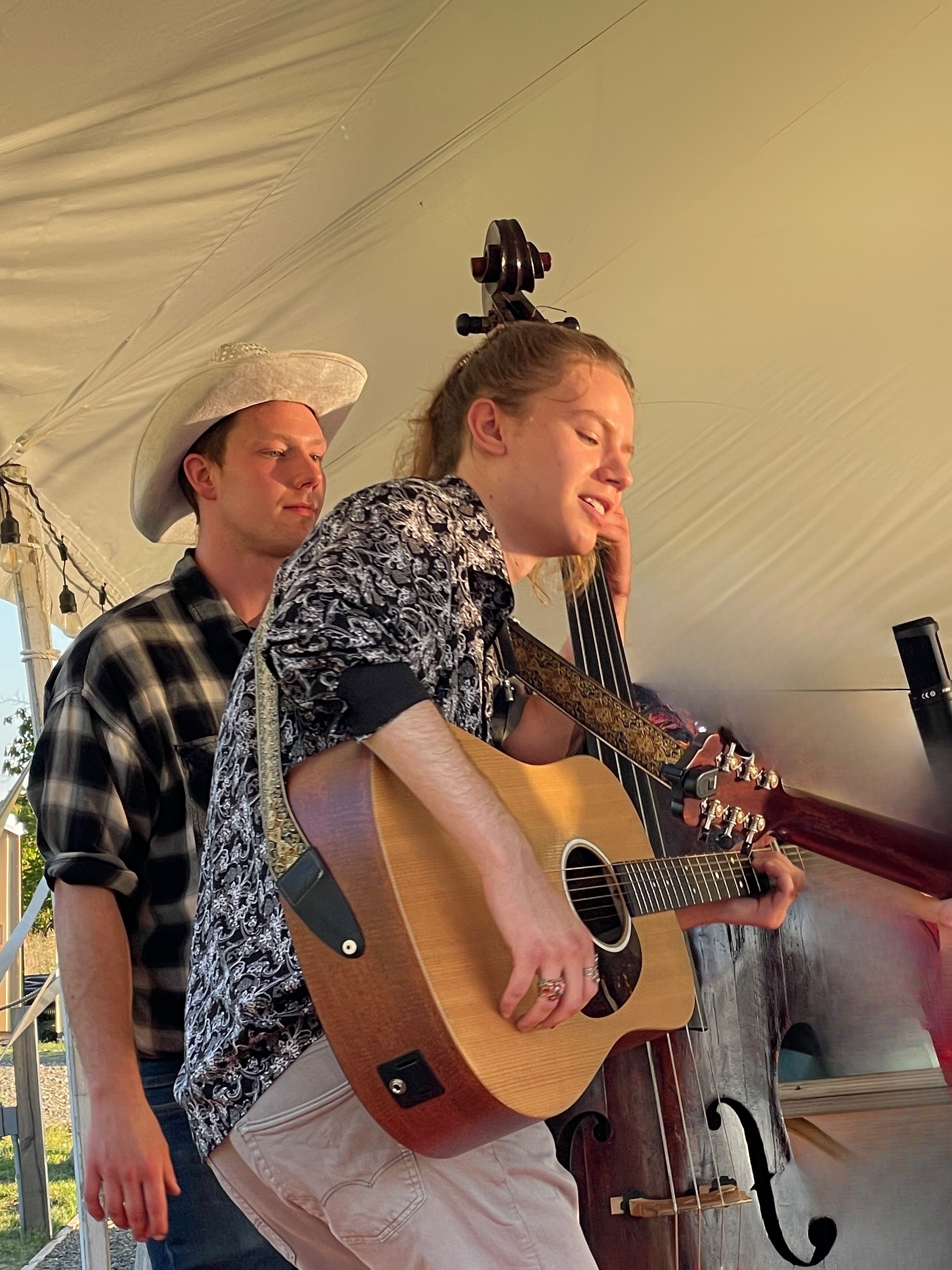 Musician singing and playing acoustic guitar onstage under a white tent, with a drummer behind her