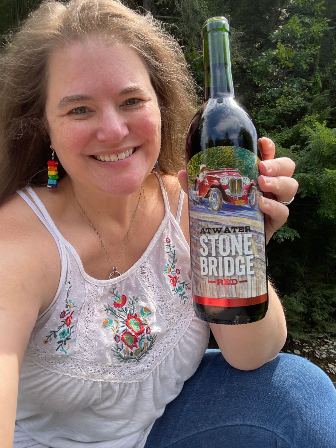 Smiling woman holding a Stone Bridge wine bottle outdoors, wearing a white embroidered top.