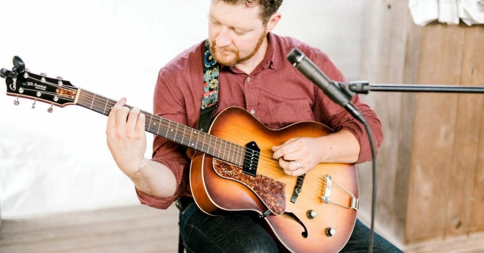 A person in a maroon shirt plays a sunburst hollow-body electric guitar near a microphone in a bright, indoor setting.