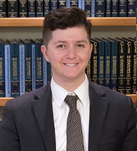 A man in a suit and tie is standing in front of a bookshelf filled with books.