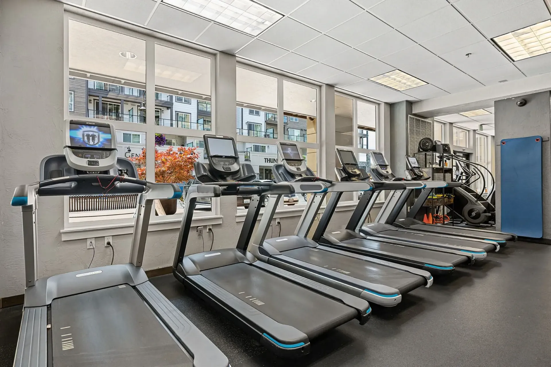 Treadmills in a fitness center with large windows overlooking apartment buildings.