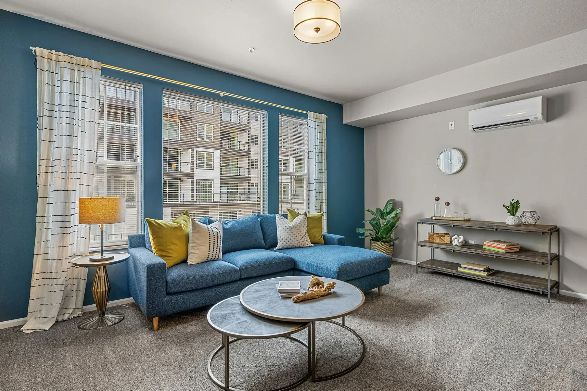 Living room with blue sectional couch, stacked coffee tables, and large windows overlooking a building.