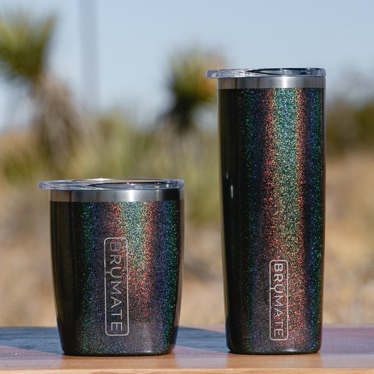 Two dark, glittery Brumate tumblers—one short, one tall—sit on a wooden surface against a blurred, sunny outdoor background.