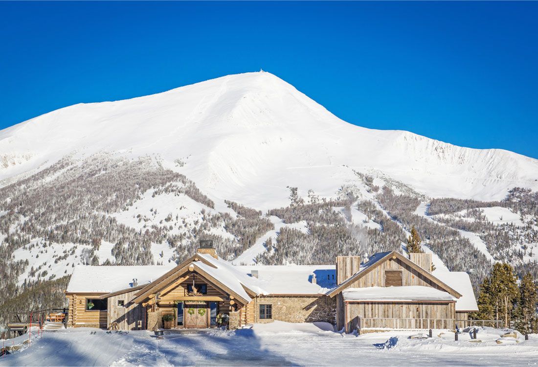 A large house is surrounded by snow with a mountain in the background.