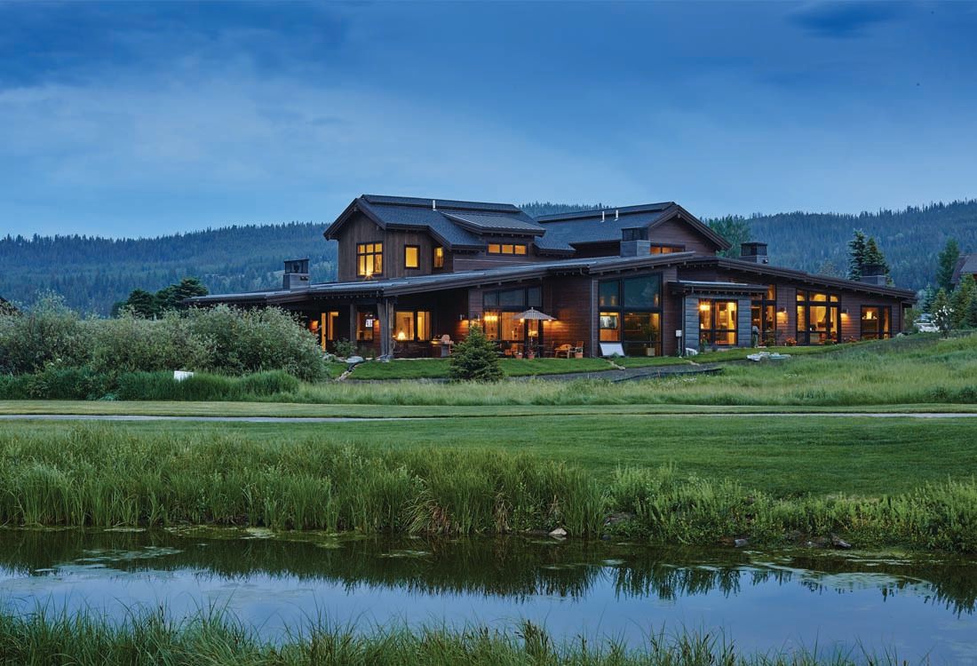 A large house is sitting in the middle of a grassy field next to a lake.