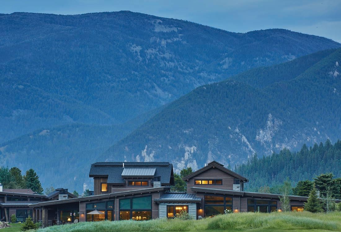 A large house is sitting on top of a grassy hill with mountains in the background.