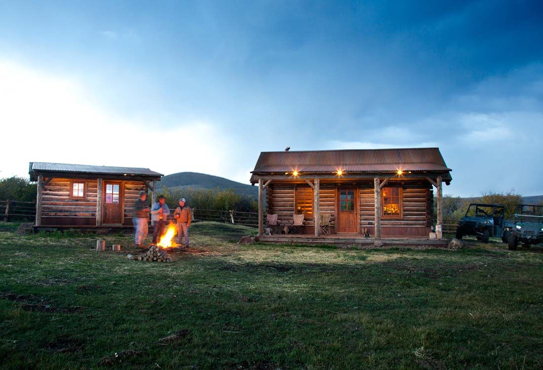 A couple is sitting around a campfire in front of a log cabin.