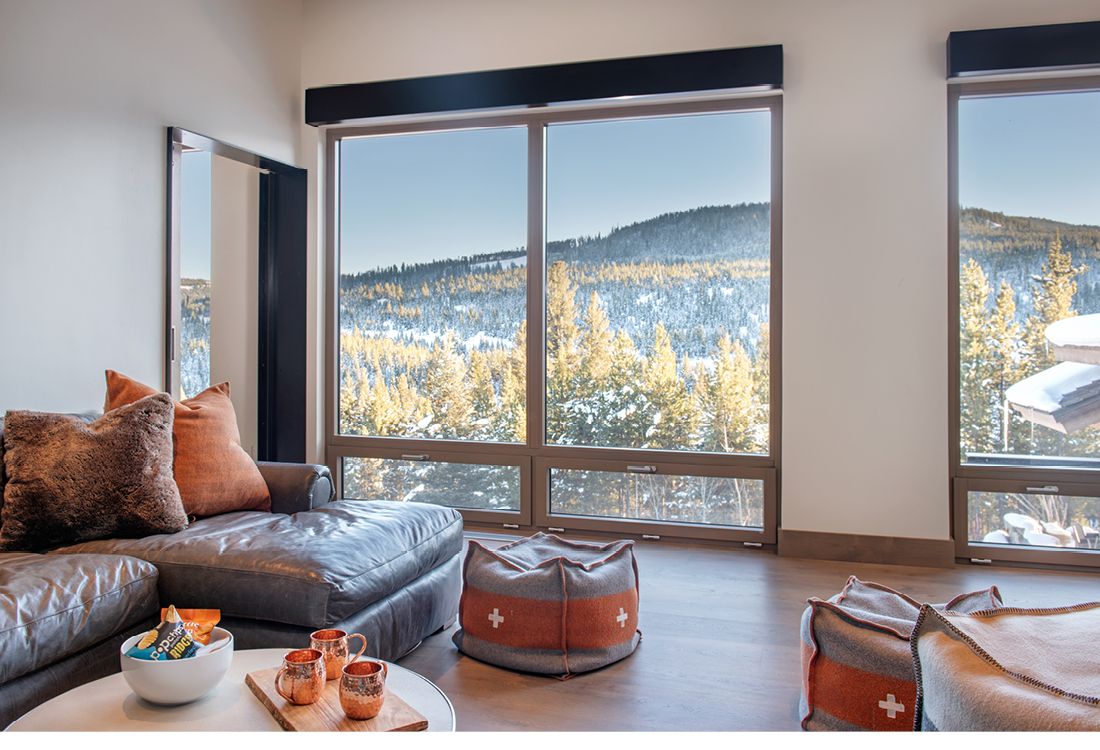 A living room with a couch , ottoman , table , and large windows.