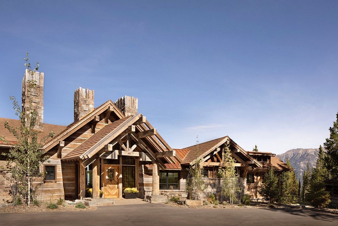 A large log cabin is sitting on top of a hill with mountains in the background.