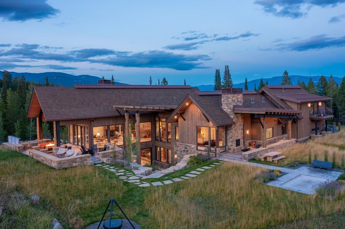 A large house is sitting in the middle of a grassy field with mountains in the background.