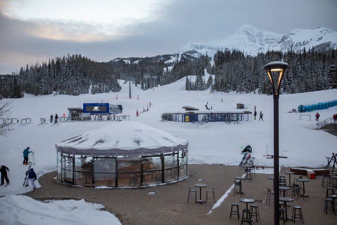 A snowy patio area with tables and chairs in front of a ski resort.