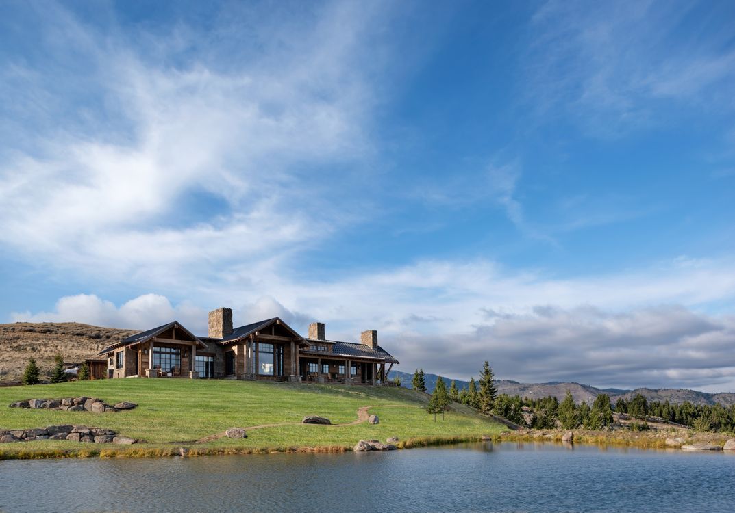 A large house is sitting on top of a grassy hill next to a lake.
