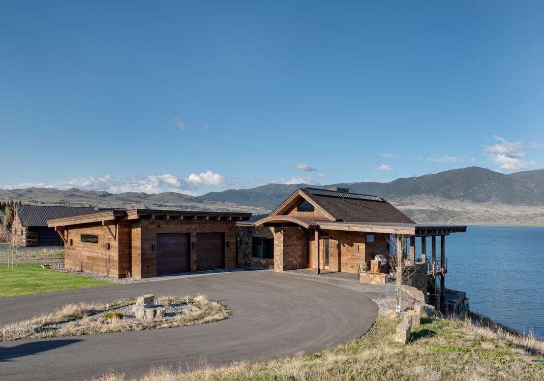 A large wooden house is sitting on top of a hill overlooking a lake.