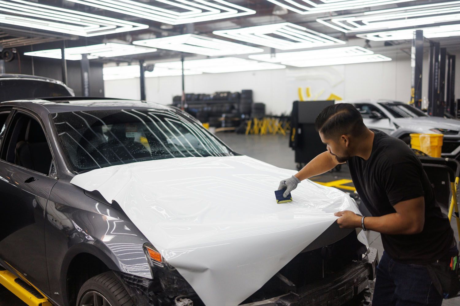 Man applying white vinyl wrap to a car hood in a garage.
