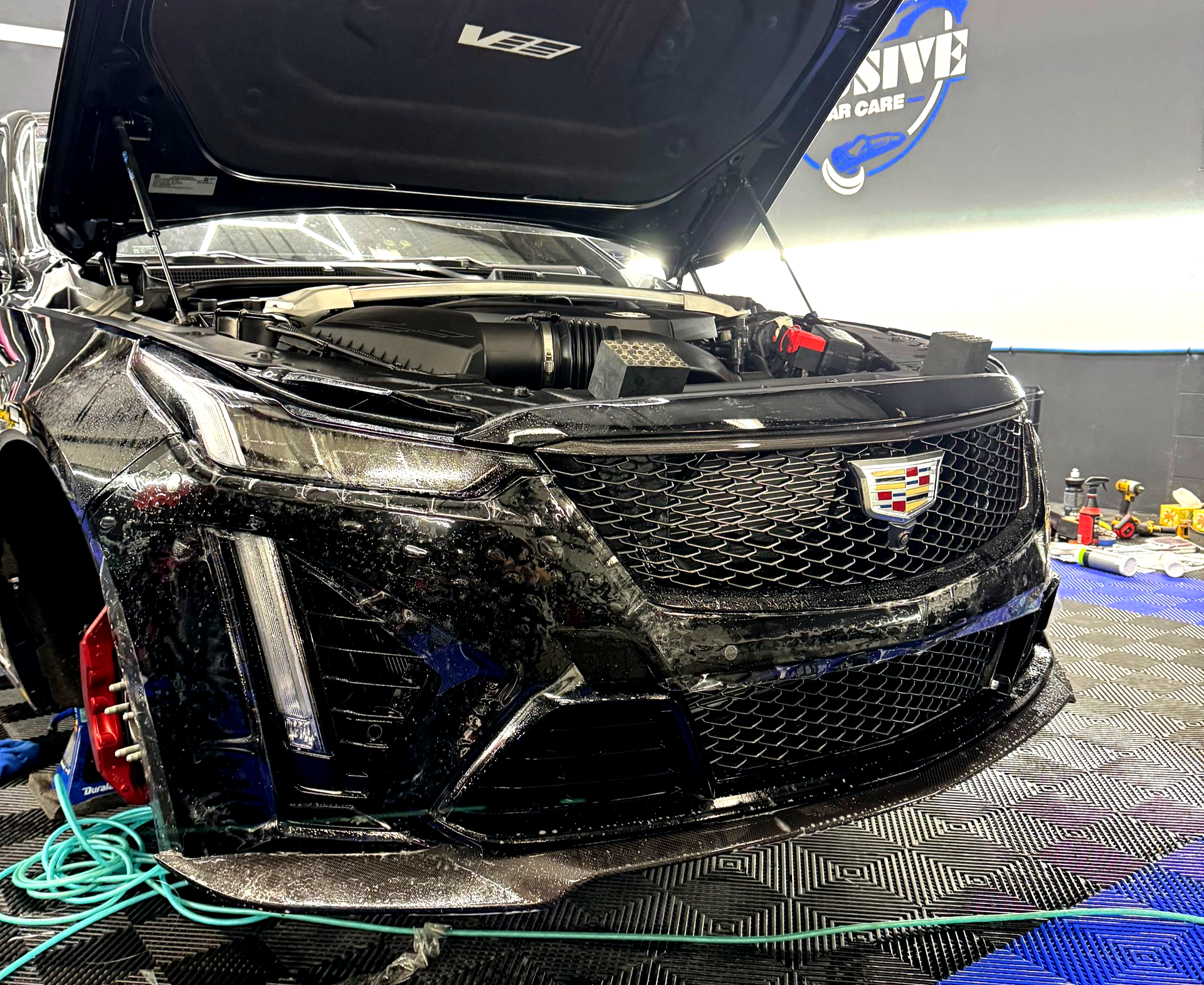 Black Cadillac with hood up, being washed on a blue tiled floor.