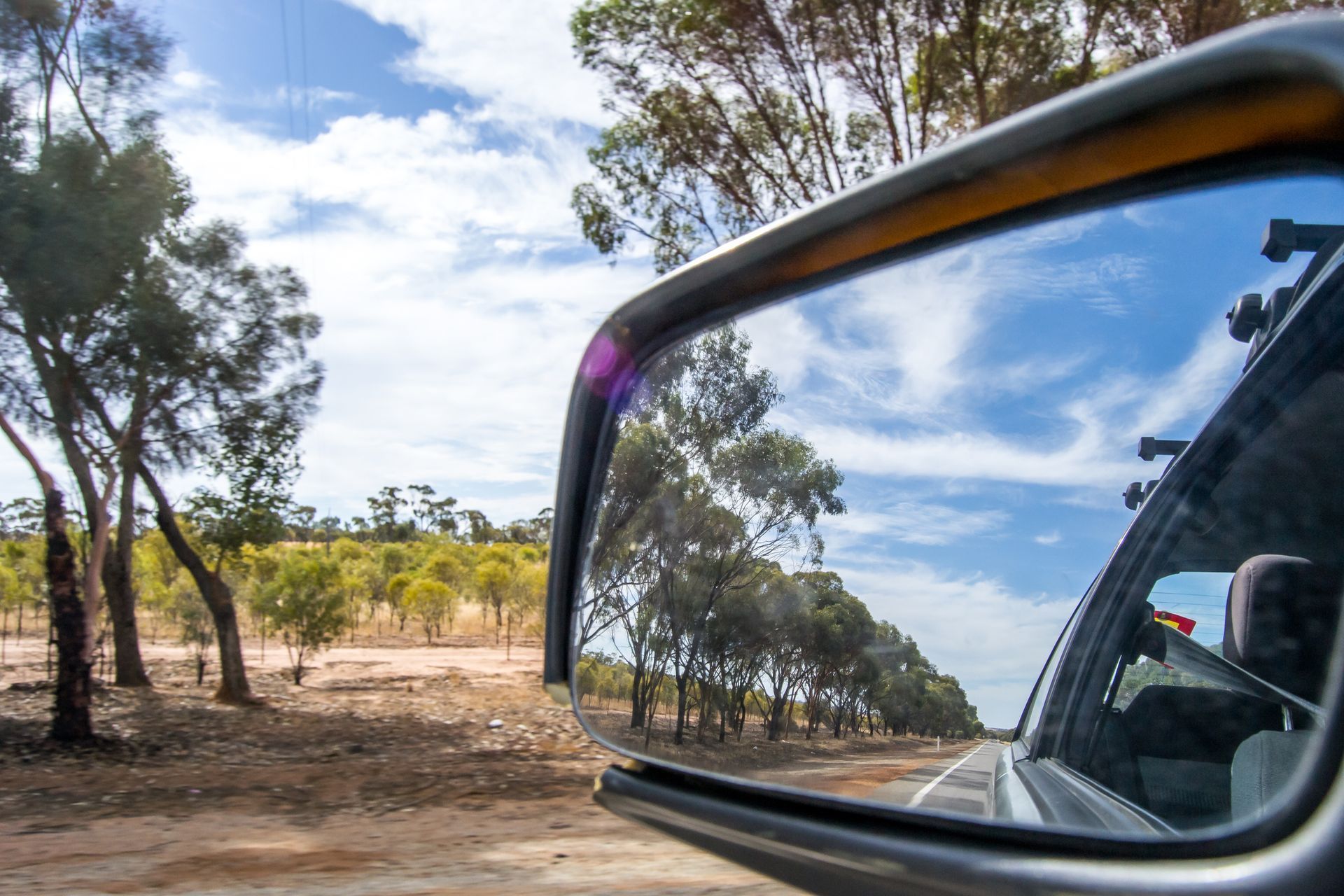 Car Side Mirror Reflecting a Tree Lined Dirt Road and Blue Sky — Eurobodalla Windscreens in Mogo, NSW