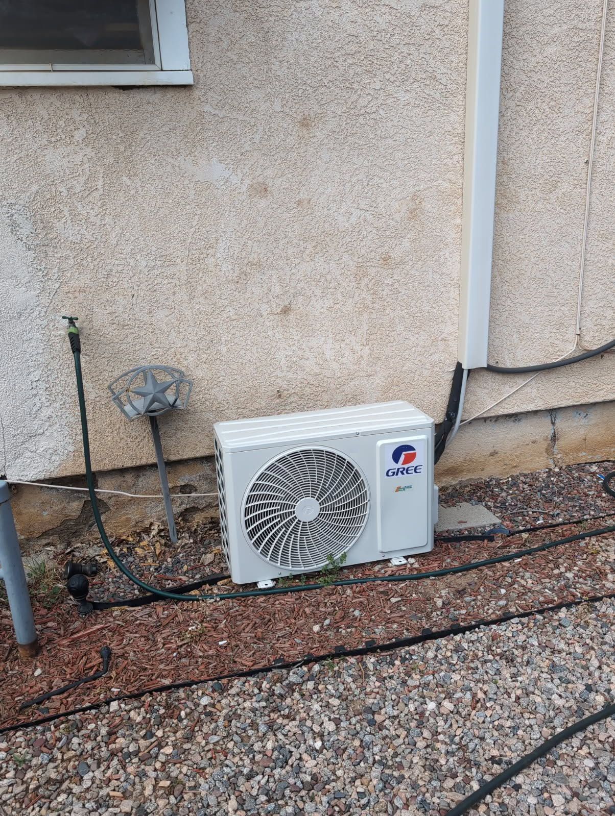 Outdoor air conditioning unit mounted against a beige wall with exposed pipes and cables on gravel ground